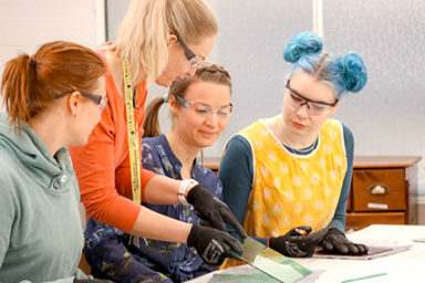 Opettaja ja opiskelijat pöydän ääressä tutkimassa lasitöiden materiaalia Käsityö- ja muotoilukoulu Näpsässä.