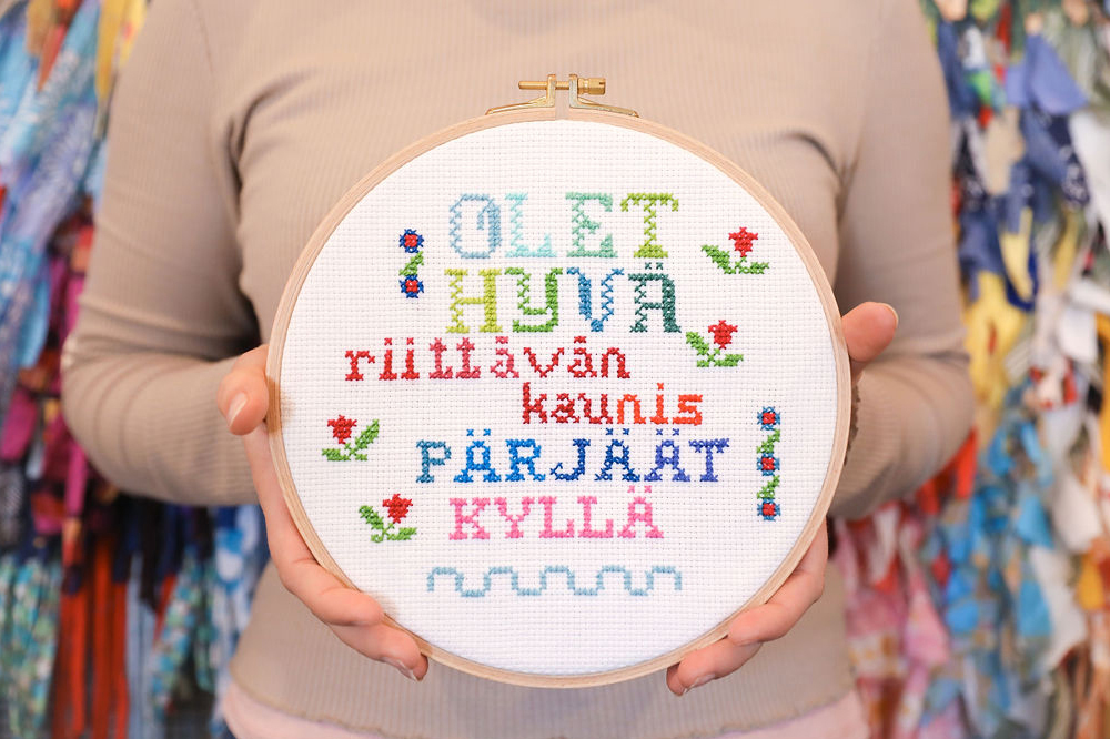 Paula Vanne pitelee kädessään kirjontakehystä. Kehykseen on kirjottu teksti "Olet hyvä, riittävän kaunis ja pärjäät kyllä".