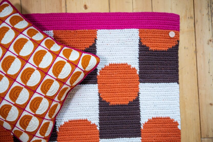 Virkattu kuviollinen matto jossa on pinkkiä, oranssia, ruskeaa ja valkoista sekä oranssivalkoinen paloista virkattu tyyny.
