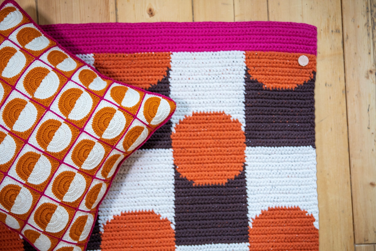 Virkattu kuviollinen matto jossa on pinkkiä, oranssia, ruskeaa ja valkoista sekä oranssivalkoinen paloista virkattu tyyny.