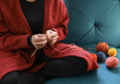 Nainen istuu sohvalla punaisessa villatakissa ja neuloo.