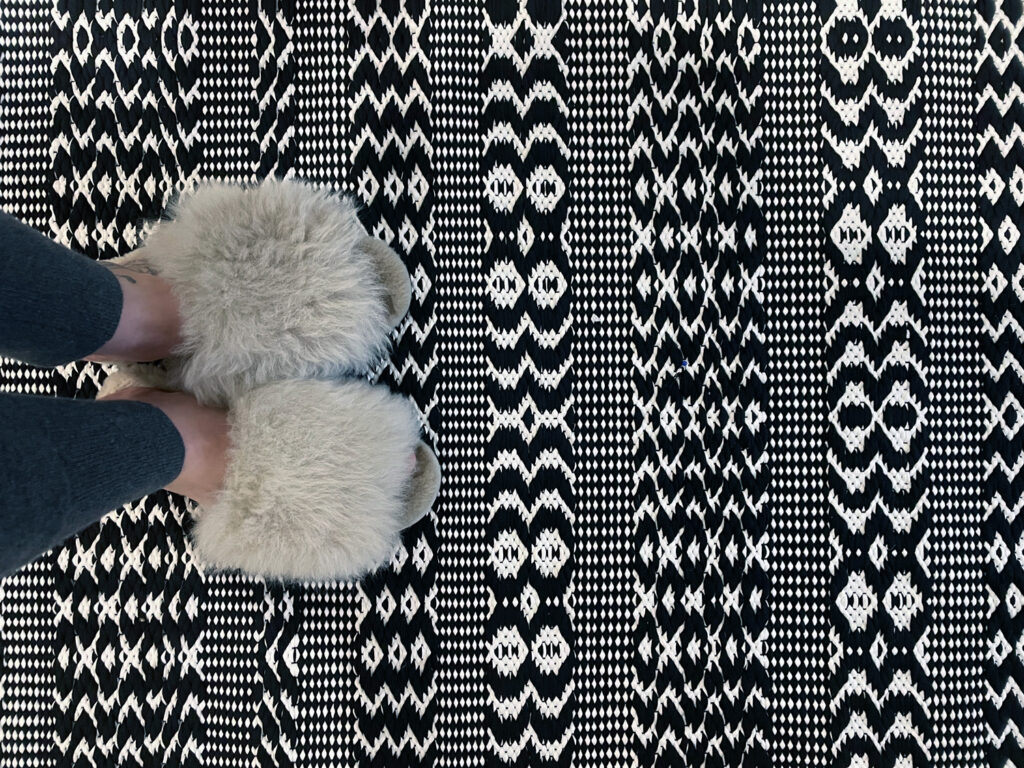 Mustavalkoinen kudottu matto, kuvassa karvatossuiset jalat maton päällä.