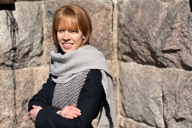 Mustatakkinen nainen kivimuurin edessä harmaasävyinen aaltotoimikashuivi kaulassaan