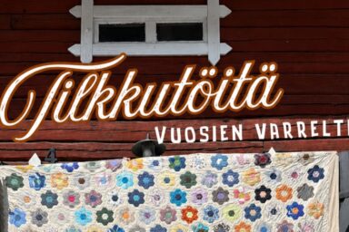 Tilkkutöitä vuosien varrelta -teksti ja kuvassa Taitokorttelin piha-aitta ja oven eteen levitetty tilkkupeitto